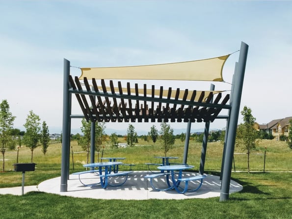 Picnic Shelter made of steel columns to shade the four picnic tables below surrounded by green grass on a sunny day