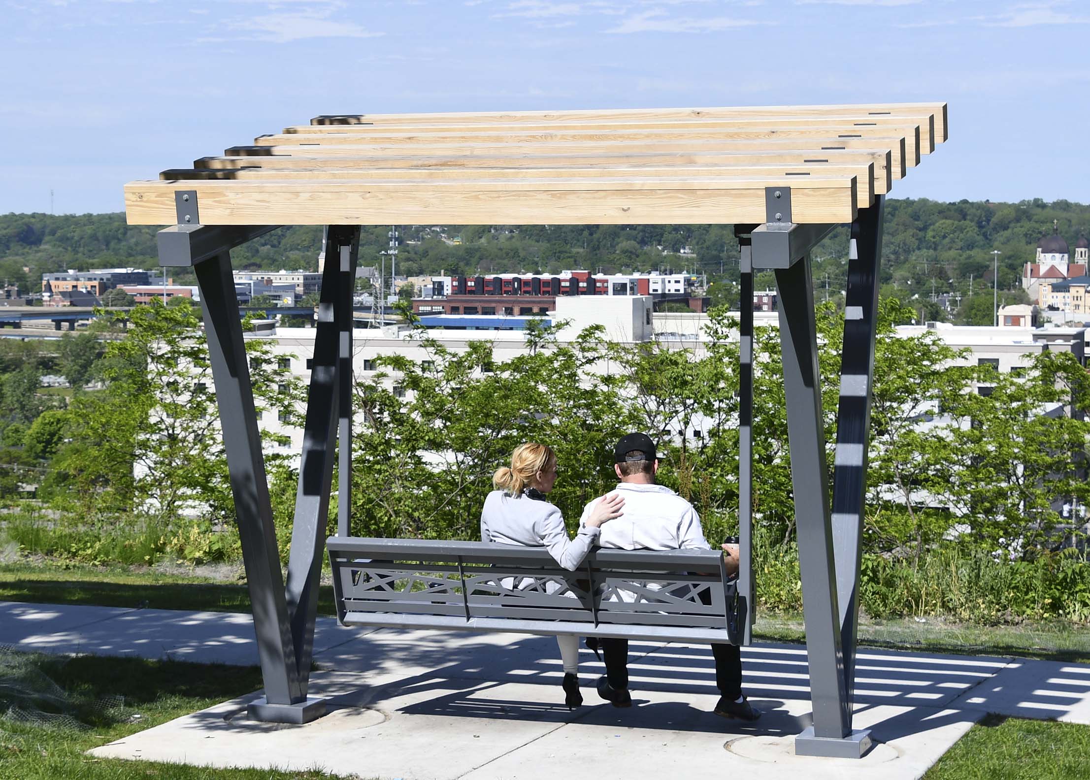 Two people sitting under the Vee Beam Swing cedar and steel structure overlooking Grand Rapids, Michigan.